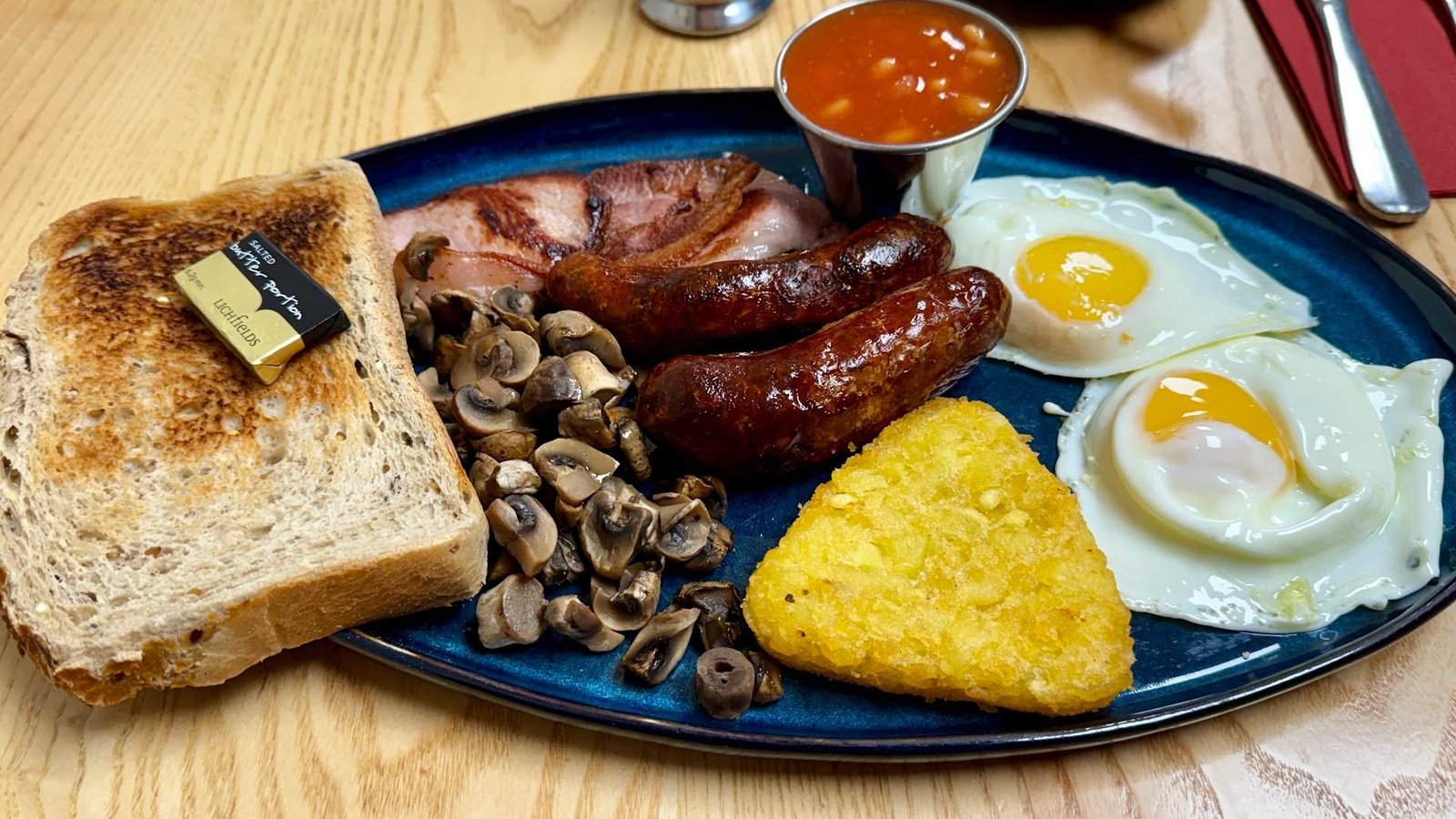 A full English breakfast on a blue plate: bacon, two sausages, mushrooms, a hash brown, two fried eggs, beans and toast with butter