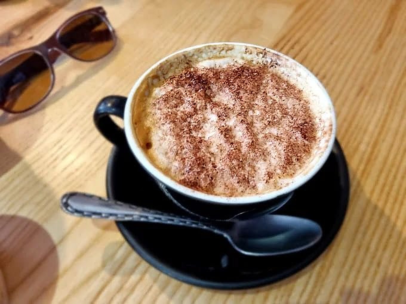 A cappuccino dusted with chocolate in a black cup and saucer on a wooden table